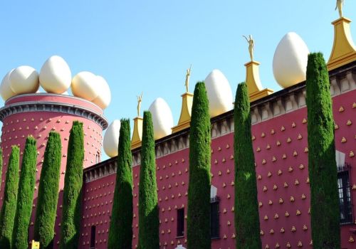 La façade rouge du Théâtre-Musée Salvador Dalí à Figueres, surmontée d'œufs géants et bordée de cyprès sous un ciel bleu à proximité du camping Les Marsouins - Ciela Village à Argelès-sur-Mer en Occitanie