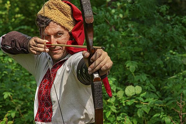 Gros plan d'un archer médiéval en costume d'époque dessinant son arc et visant avec une flèche au Parc Durandal à proximité du camping Les Cigales - Ciela Village dans le Lot